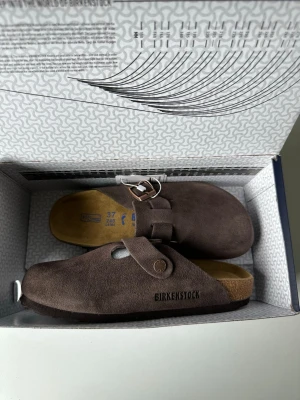 Birkenstock Boston mörkbruna sandaler - Mörkbruna Birkenstock Boston sandaler i mocka med justerbart spänne och klassisk korksula. Perfekta för en avslappnad och trendig stil. Modellen har stängd tå och bekväm fotbädd som formar sig efter foten.
