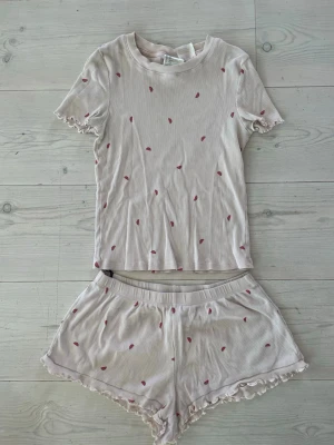Ljusrosa pyjamasset med vattenmelonstryck - Sött pyjamasset i ljusrosa med små vattenmeloner som tryck. Setet består av en T-shirt  och shorts. Mjukt och stretchigt material 