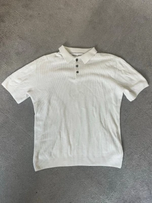 Vit stickad polotopp med knappar - Snygg vit stickad polotopp med korta ärmar och tre knappar framtill från Jack & jones. Toppen har en klassisk krage och ett diskret, strukturerat mönster i materialet som ger en modern touch. Perfekt för dig som gillar stilrena och enkla plagg.