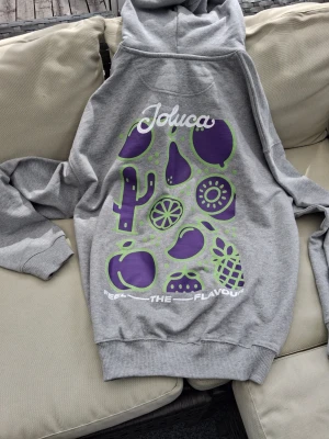 Grå hoodie med fruktmotiv från Joluca - Snygg grå hoodie från Joluca med stor färgglad print på ryggen i lila och grönt med frukter och kaktus. Framtill finns en liten logga på bröstet. Klassisk huva och ribbade muddar. Perfekt för dig som gillar streetwear och unika prints. Limited edition