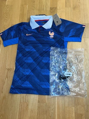 Blå Nike Frankrike fotbollströja - Snygg blå fotbollströja från Nike med Frankrikes landslagslogga på bröstet. Tröjan har ett mönster i olika blå nyanser, vit krage och orange Nike-logga. Perfekt för dig som älskar fotboll och vill visa ditt stöd för Frankrike. 
