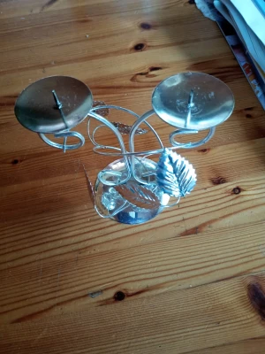 Silvrig ljusstake med lövdetaljer - Säljer en silvrig ljusstake i metall med plats för två ljus. Ljusstaken har dekorativa lövdetaljer och en rund fot. Perfekt som snygg inredningsdetalj på bordet eller i fönstret.