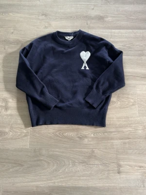 Mörkblå Ami Paris tröja med hjärtdetalj - Mörkblå stickad tröja från Ami Paris med rund halsringning och ribbade muddar. På bröstet finns ett stort vitt hjärta med bokstaven A. Perfekt för dig som gillar stilrena och trendiga plagg med en twist.