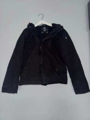 Svart vindjacka från Rip curl - Svart vindjacka från Rip Curl med huva och dragkedja framtill. Jackan har två stora fickor med lock och knapp, samt diskret logga på ärmen. Perfekt för blåsiga dagar och enkel att matcha med olika outfits.