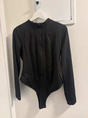 Svart långärmad body med dragkedja - Snygg svart body med långa ärmar och dragkedja framtill. Tight passform och rund halsringning. Perfekt att styla till jeans eller kjol. Materialet känns stretchigt och följsamt. Enkel och stilren design utan synliga loggor.