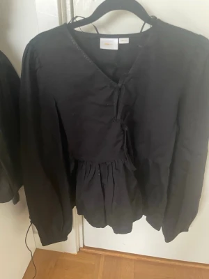 Svart blus från Gina Tricot - Svart blus från Gina Tricot med v-ringning och knytning framtill. Blusen har långa ärmar och en lätt rynkad midja som ger en snygg siluett. Perfekt att styla med jeans eller kjol för en trendig look. Den ser dock skrynklig ut men den har kommit från tvätten och ska strykas inom kort🌺❣️☀️