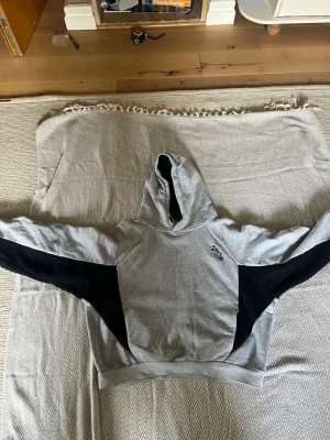Grå och svart bigstep hoodie från Mertra - Snygg hoodie i grått med svarta paneler på ärmarna från Mertra. Hoodien har broderad logga på bröstet och en klassisk huva. Perfekt för en avslappnad streetwear-look. Mjuk bomullsblandning och ribbade muddar.storlek s passar även xs