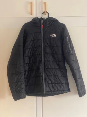 Svart pufferjacka från The North Face - Svart pufferjacka från The North Face med huva och hel dragkedja framtill. Jackan har quiltad design, logga på bröstet och är tillverkad i ett lätt, syntetiskt material. Perfekt för kalla vinterdagar.