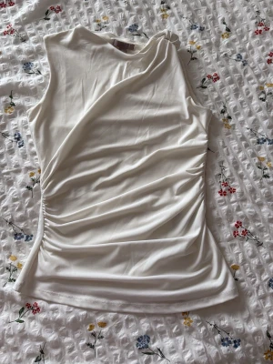Vit ärmlös topp från H&M - Snygg vit ärmlös topp från H&M med draperad effekt på ena sidan. Toppen har en rund halsringning och är tillverkad i ett mjukt, stretchigt material som sitter bekvämt och smickrande. Perfekt att styla med jeans eller kjol. Small defekt på se gärna bild.