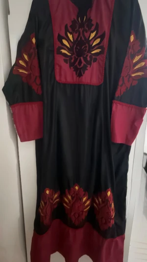 Svart och vinröd klänning med broderi - Unik långklänning i svart och vinrött med vackra broderade detaljer i svart och gult på bröst, ärmar och nederkant. Klänningen har långa ärmar och en lös passform, perfekt för dig som gillar bohemiska och färgstarka plagg.