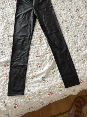 Svarta skintight leggings i fuskskinn  - Snygga svarta leggings i fuskskinn med hög midja och tight passform. De har en slät yta och är perfekta för att skapa en trendig look. Materialet är glansigt och stretchigt för extra komfort.
