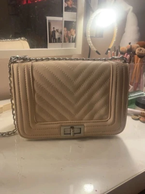 Beige quiltad handväska med kedja - Säljer en beige quiltad handväska med silverfärgad kedjerem och snyggt mönster i v-form. Väskan har ett metallspänne framtill och är tillverkad i konstläder. Perfekt för dig som gillar stilrena accessoarer med trendig touch.