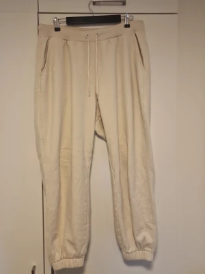 Beige mjukisbyxor med dragsko - Beiga mjukisbyxor med resår i midjan och benslut samt dragsko för perfekt passform. Byxorna har två sidofickor och är tillverkade i ett mjukt och bekvämt material, perfekta för chill dagar. Enkel och stilren design som passar till det mesta.