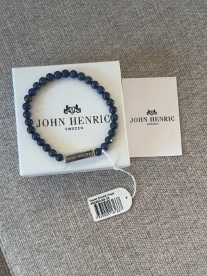 Blått pärlarmband från John Henric - Stilrent armband med blå pärlor och en silverfärgad metallbricka med ingraverad logga från John Henric. Elastiskt band gör det enkelt att ta på och av. Perfekt accessoar för att lyfta din outfit.