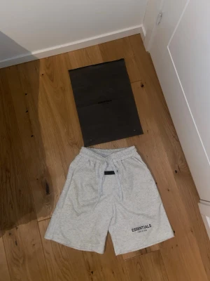Essentials Shorts - Snygga ljusgrå shorts från Essentials Fear of God med elastisk midja och snörning. Mjukt bomullsmaterial och logga tryckt på ena benet. Perfekta för en avslappnad och trendig stil.