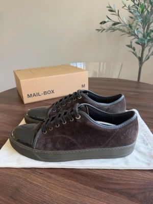 Lanvin - Snygga bruna sneakers med ovandel i mjuk mocka och tåparti i blankt lack. Skorna har matchande bruna skosnören och en robust sula i mörkgrått. Perfekta för dig som gillar en stilren och modern look med en twist av olika material.