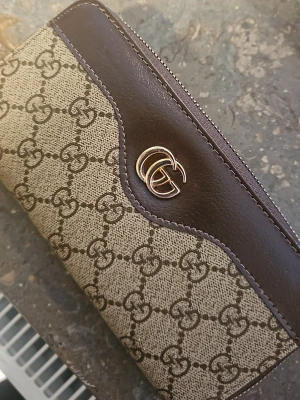 Brun plånbok med GG-mönster - Snygg plånbok i brunt läder och canvas med klassiskt GG-mönster. Plånboken har guldfärgad logga, dragkedja runtom och flera fack för kort och mynt. Perfekt för dig som gillar lyxiga accessoarer och vill ha ordning på dina grejer.