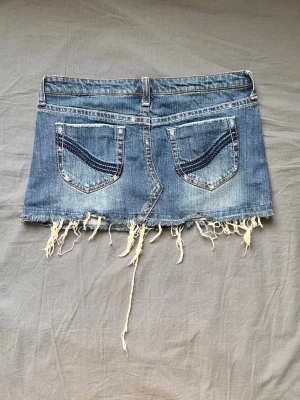 Blå jeanskjol med fransig kant - Kort jeanskjol från Only i klassisk blå denim med slitna detaljer och fransig, rå kant nedtill. Kjolen har fem fickor, bälteshällor och knappstängning fram. Perfekt för dig som gillar en avslappnad och trendig stil. Midjemått 76cm