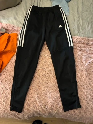 Svarta Adidas träningsbyxor - Svarta träningsbyxor från Adidas med klassiska vita ränder längs sidorna och Adidas-logga på vänster lår. Byxorna har smal passform, resår i midjan och är tillverkade i ett lätt och stretchigt material som passar perfekt för sport och träning.