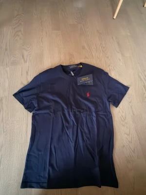 Mörkblå t-shirt från Polo Ralph Lauren - Snygg mörkblå t-shirt från Polo Ralph Lauren med klassisk rund hals och ikoniska röda broderade loggan på bröstet. Tillverkad i mjuk bomull som känns skön mot huden. Perfekt för en clean och stilren look. 100% bomull