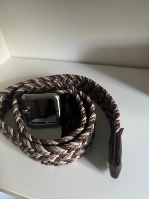  flätat bälte - Snyggt flätat bälte i bruna och beige toner med silverfärgat spänne. Bältet har detaljer i skinn vid spännet och änden, vilket ger en stilren och trendig look. Perfekt accessoar för att lyfta din outfit.