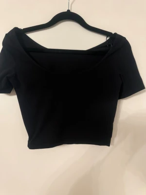 Svart croppad topp med bred urringning - En svart croppad topp med bred urringning och korta ärmar. Snygg och enkel design som passar perfekt till jeans eller kjol. Toppens material känns mjukt och stretchigt, vilket gör den bekväm att bära hela dagen.