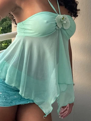 Mintgrön omlott topp med blomdetalj - Superfin mintgrön topp med omlottkänsla och asymmetrisk nederkant. Toppen är ärmlös med tunna axelband och har en stor vit blomma framtill som detalj. Luftigt material som ger en flowy look, perfekt för varma dagar.