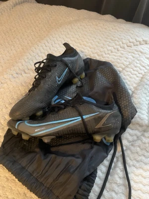 Nike Mercurial svarta fotbollsskor - Säljer ett par svarta Nike Mercurial fotbollsskor med ljusblå detaljer och snörning. Skorna har en modern, aerodynamisk design och är tillverkade i syntetmaterial för bästa grepp och känsla på planen. Perfekta för dig som vill ha stilrena och snabba skor.