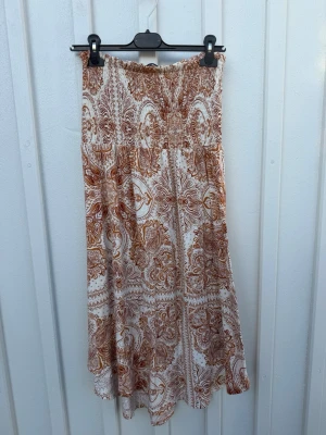 Tube sommarklänning!  - Axelbandslös midiklänning i varma toner av brun och beige. Smockad överdel som sitter bekvämt och fint över bysten samt en luftig, flowy passform som faller mjukt. Perfekt sommarklänning för semester, stranddagar eller avslappnade sommarkvällar. 🧡🤎
