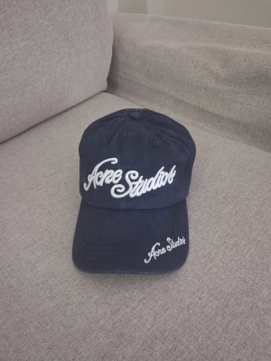 Mörkblå keps från Acne Studios - Snygg mörkblå keps från Acne Studios med vit broderad logga framtill och på skärmen. Justerbar rem baktill med metallspänne. Klassisk böjd skärm och stilren design, perfekt för en trendig streetwear-look.
