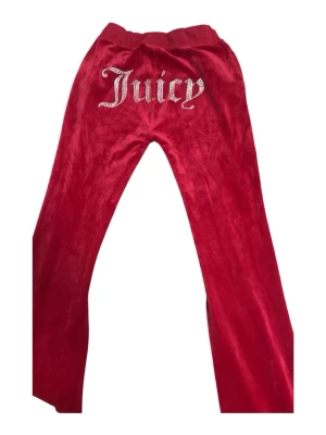 Röda velourbyxor från Juicy Couture - Röda mjukisbyxor i velour från Juicy Couture med strassdetaljer och texten 'Juicy' på baksidan. Byxorna har utsvängda ben, dragsko i midjan och broderad logga fram. Perfekta för en trendig och avslappnad look.