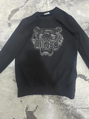 Svart sweatshirt med tigertryck Kenzo - Svart sweatshirt från Kenzo med ikoniskt tigertryck och logga framtill. Tröjan har rund halsringning, långa ärmar och ribbade muddar vid ärmslut och nederkant. Perfekt för en trendig och avslappnad stil.
