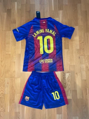 FC Barcelona matchställ Lamine Yamal - Säljer ett FC Barcelona matchställ med Lamine Yamal och nummer 10. Setet består av en blå och röd randig tröja med klubbmärke och sponsorlogga samt matchande shorts med klubbmärke och nummer. Perfekt för fotbollsträning eller att bära som supporter.