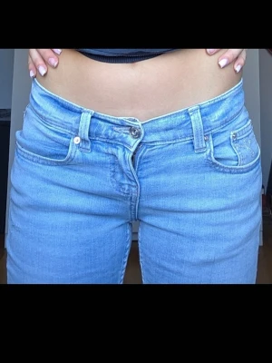 Ljusblå bootcut jeans med slitningar - Säljer ett par ljusblå bootcut jeans med slitningar på låret och knät. Jeansen har klassisk femficksdesign, hällor för skärp och stängs med knapp och dragkedja. Perfekta för en avslappnad och trendig look.