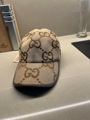 Äkta  keps från Gucci säljer den billigt för jag hat tappat bort kvittot  - Säljer en  keps från Gucci med det klassiska GG-mönstret i brunt. Kepsen är tillverkad i canvasmaterial och har en böjd skärm. Märket syns tydligt både på utsidan och på etiketten inuti kepsen. Perfekt accessoar för att lyfta din stil.