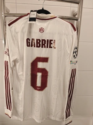 Arsenal spelar version långärmad matchtröja Gabriel - Säljer en vit långärmad Arsenal spelar Version matchtröja från Adidas med vinröda detaljer och tryck. Tröjan har namn och nummer 'Gabriel 6' på ryggen, Champions League-märke på ärmen och klassisk krage. Materialet är lätt och andas, perfekt för fotbollsträning eller match.