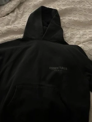 Svart Essentials Fear of God hoodie -  Intresse koll på en Svart hoodie från Essentials Fear of God med diskret logga framtill och stor logga på ryggen. Klassisk känguruficka och med huva.