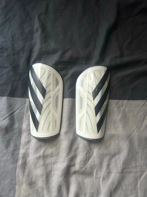 Vita benskydd från Adidas - Säljer ett par vita benskydd från Adidas med svarta ränder. De har en lätt och smidig form som skyddar smalbenen vid fotbollsspel. Tillverkade i syntetmaterial och designade för att sitta bekvämt under strumpan.