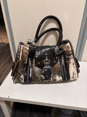 Y2K väska  - Säljer en unik handväska från LYDC med ormmönster i beige, brunt och svart. Väskan har svarta detaljer, dubbla flätade handtag, dekorativa dragkedjor och en metallkrona på framsidan. Snygga nitar och spännen ger en edgy look. Perfekt för dig som gillar statement-accessoarer. Säljer pågrund av att väskan har tappat färg. Den kan lämna färg smulor efter sig 