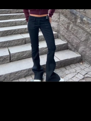 Svarta bootcut jeans med knappar - Säljer ett par svarta bootcut jeans med två vita knappar i midjan och fickor fram. Jeansen har utsvängda ben och sitter tight upptill för en snygg siluett. Perfekta till sneakers eller boots för en trendig  look.
