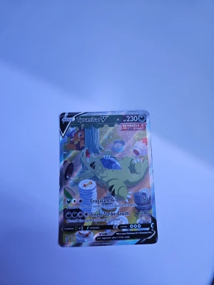 Tyranitar V Full Art - Säljer ett Pokémonkort av typen Tyranitar V Full Art från Sword & Shield-serien. Kortet har en färgglad och glansig design med Tyranitar som ligger under ett träd omgiven av mat och burkar. Perfekt för samlare eller Pokémonfans. 