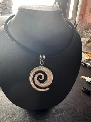 Halsband med spiralformad silverberlock - Unikt halsband med en stor spiralformad berlock i silverfärgad metall. Hängs på ett svart, tjockt band som ger en cool kontrast till den glänsande spiralen. Perfekt för dig som gillar statement-smycken och vill sticka ut med en modern och konstnärlig look.