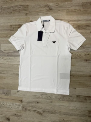 Vit pikétröja från Prada - Snygg vit pikétröja från Prada med klassisk krage och korta ärmar. Tröjan har en liten svart logotyp på bröstet och knäppning framtill. Perfekt för en stilren och clean look.