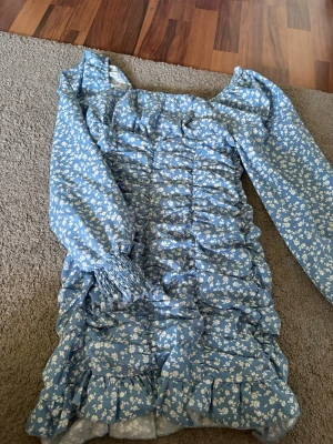 Blå blommig kortklänning med volang - Supersöt blå kortklänning med vita blommor och volangdetaljer. Klänningen har en dragkedja på sidan, långa ärmar med smock vid ärmslut och rynkade partier framtill. Perfekt för vår och sommar, ger en romantisk och trendig look.