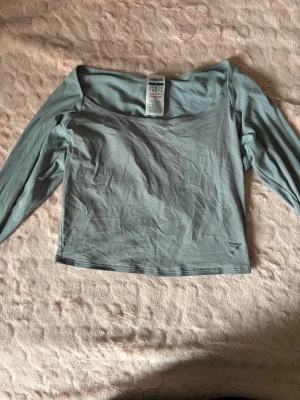 Ljusgrön långärmad offshoulder topp - Trendig ljusgrå offshoulder topp med långa ärmar från gymshark. Toppens material är mjukt och stretchigt, perfekt för en cool och avslappnad stil. Enkel design utan mönster eller detaljer, vilket gör den lätt att matcha med olika outfits.
