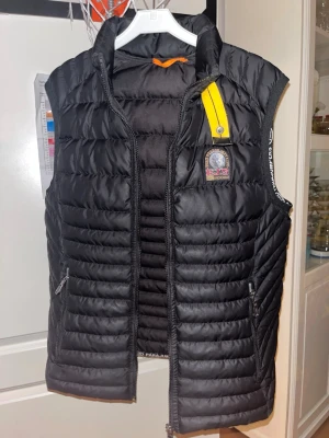 Svart dunväst från Parajumpers - PASSAR S, PRIS KAN DISKUTERAS. Svart quiltad dunväst från Parajumpers med gul detalj vid kragen och logotyp på bröstet. Västen har dragkedja framtill och två fickor med dragkedja. Perfekt för lager-på-lager och snygg till många outfits.