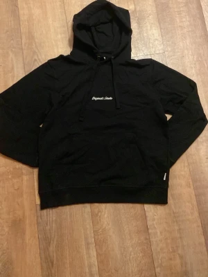 Svart hoodie från Jack and Jones - Svart hoodie från Jack and Jones med vit text på bröstet. Klassisk modell med huva och dragsnören samt en stor magficka framtill. Perfekt för en avslappnad och stilren look. Priset är inte hugget i sten och hör gärna av er vid frågor!