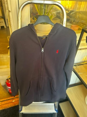 Mörkblå hoodie från Polo Ralph Lauren - Snygg mörkblå hoodie med dragkedja från Polo Ralph Lauren. Klassisk design med röd logga broderad på bröstet, huva och kängurufickor framtill. Tillverkad i mjukt bomullsmaterial som är skönt att ha på sig.