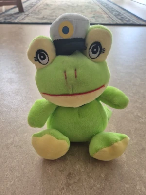 Mjukisdjur - Söt grön groda som mjukisdjur med stor studentmössa och en gul-blå scarf runt halsen. Grodan har stora ögon med långa ögonfransar och ljusgula fötter. Perfekt som present till studenten eller som dekoration. Tillverkad i mjukt plyschmaterial.