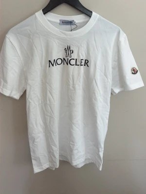 Vit Moncler t-shirt med logga - Snygg vit t-shirt från Moncler med klassisk svart logotyp på bröstet och Moncler-märke på ärmen. T-shirten har rund hals och korta ärmar, perfekt för dig som gillar stilrena plagg med exklusiv känsla. Materialet är mjuk bomull.